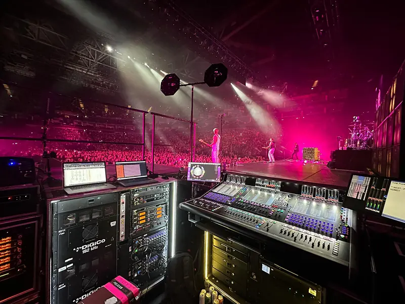 Front of House position with a DiGiCo console, racks and laptops beside a pink-lit arena stage with the band performing