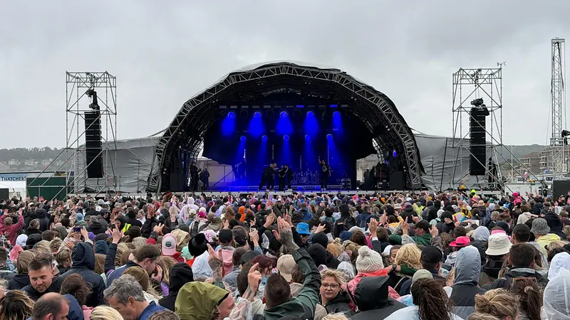 Outdoor festival stage under a grey sky with blue stage lighting and a crowd in rain ponchos