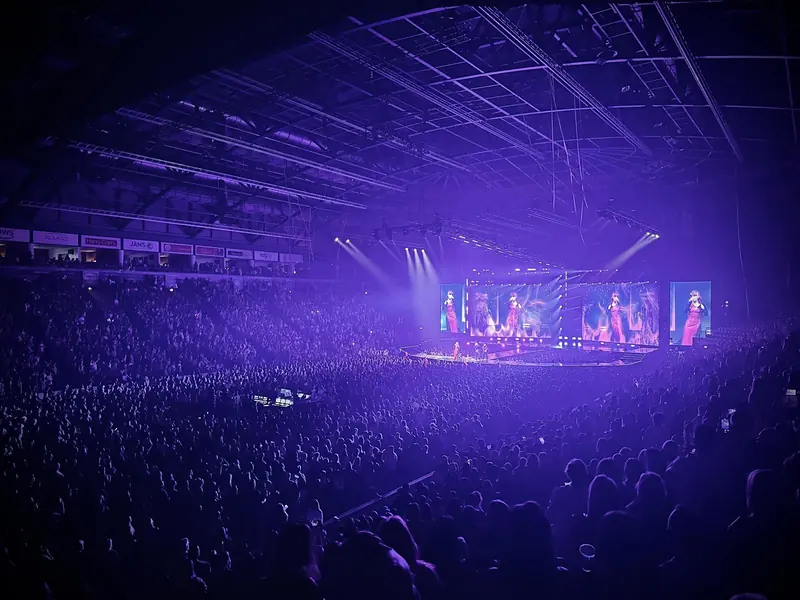 Wide view of a packed arena in purple light with large video screens either side of the stage